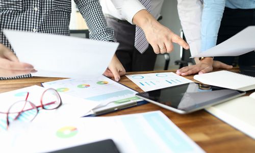 Close-up of people hands and important business paper lying on wooden table. Female hand holding sheet of paper. Man standing and pointing finger on tablet with empty screen